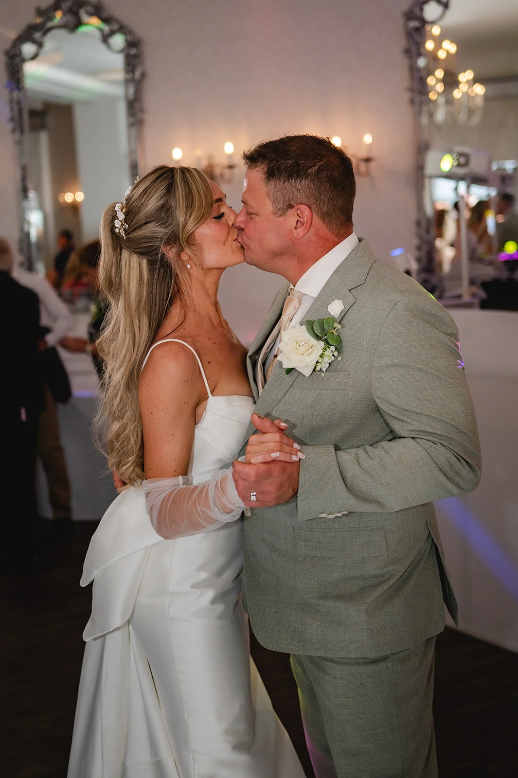A bride and groom share a tender kiss during their first dance, with the bride wearing a sleek satin gown with sheer gloves and the groom in a smart sage suit.