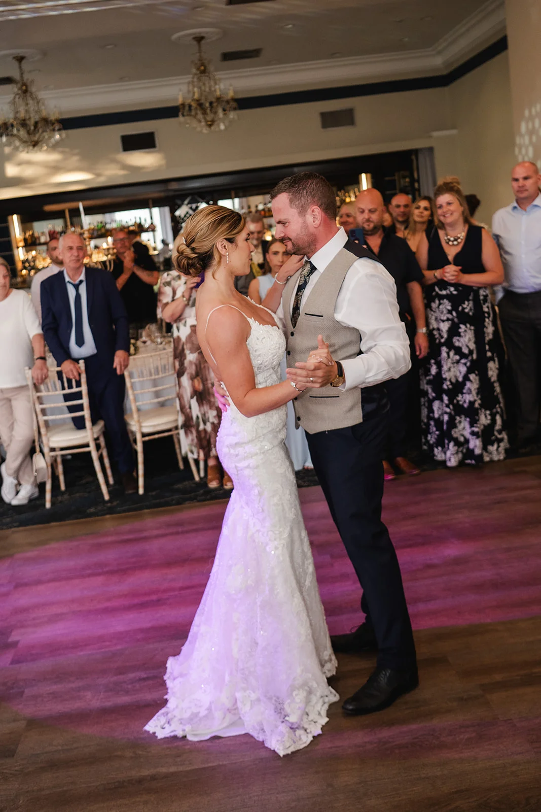 The newly married couple share an intimate first dance on a wooden dance floor surrounded by watching family and friends in a brightly lit hotel ballroom.