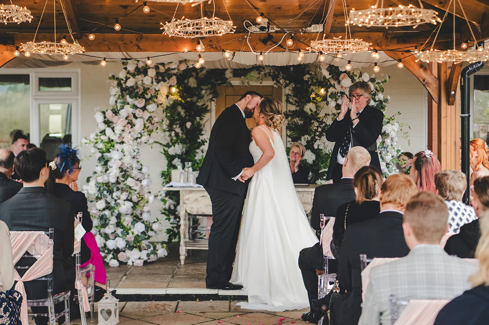 Newlywed bride and groom share their first kiss under a floral arch during an outdoor ceremony while guests applaud.