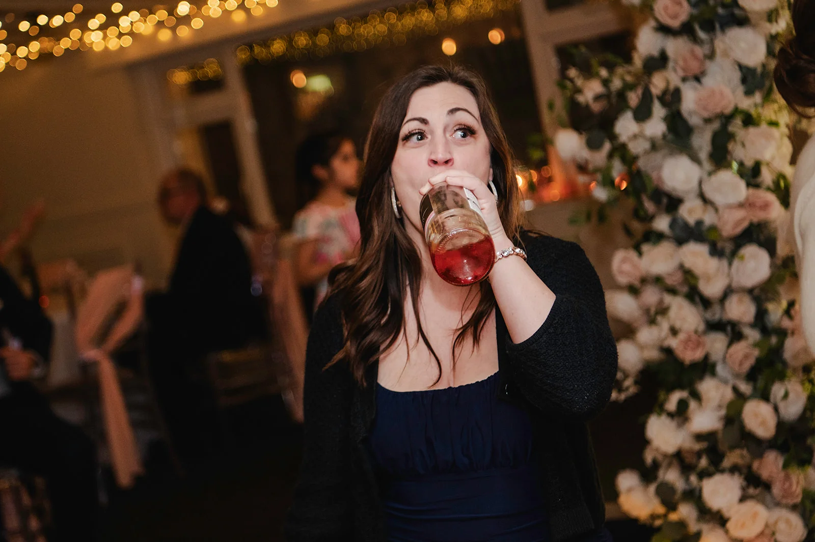 A wedding guest takes a humorous swig from a bottle of drink next to a large floral arrangement and warm fairy lights during the reception.
