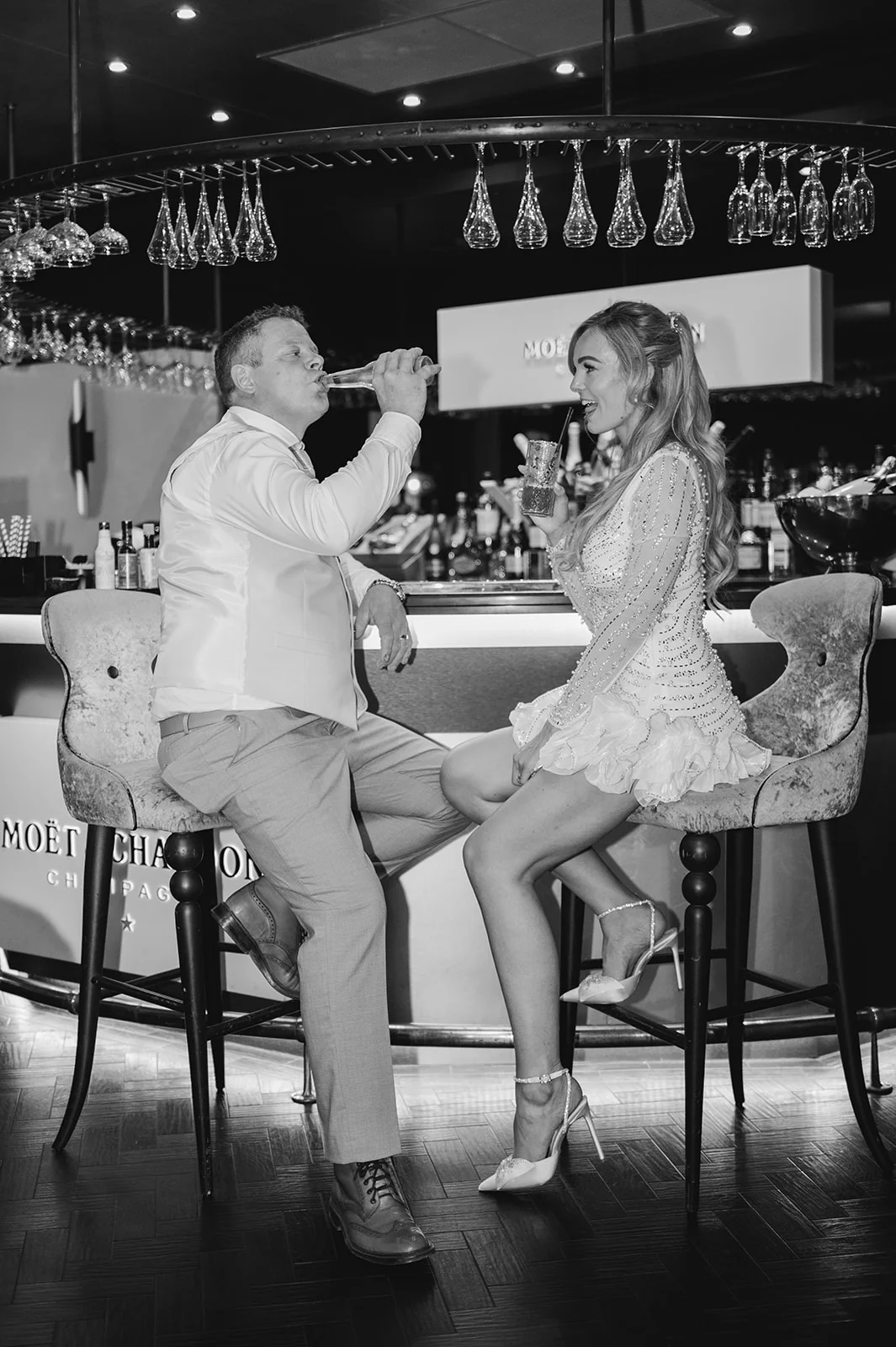 A newlywed couple sits on bar stools at a hotel bar during their evening reception, sharing a joyful laugh while enjoying drinks together.