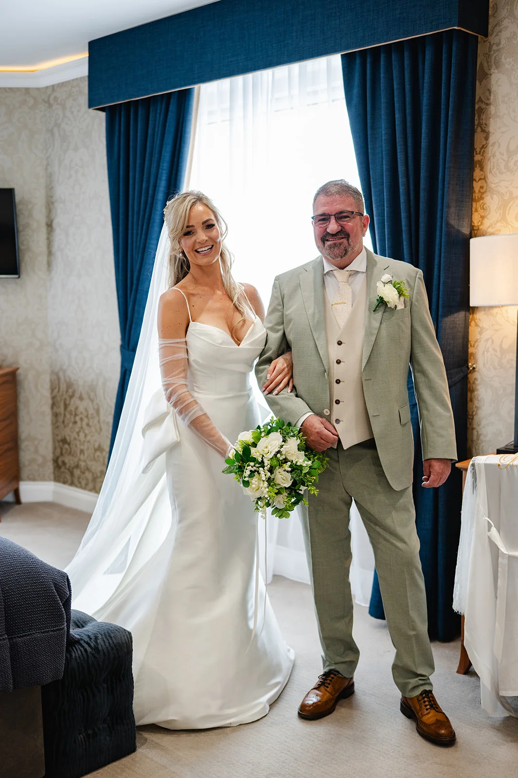 A smiling bride in a sleek white satin gown with sheer sleeves and a veil stands alongside her father in a sage green suit holding a bouquet of white roses.