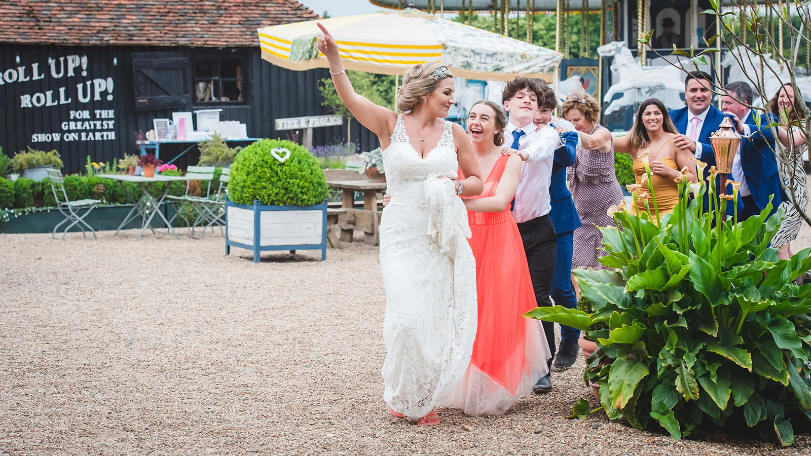 A joyful bride in a lace gown and tiara leads a lively group of laughing wedding guests outdoors near a vintage funfair carousel.