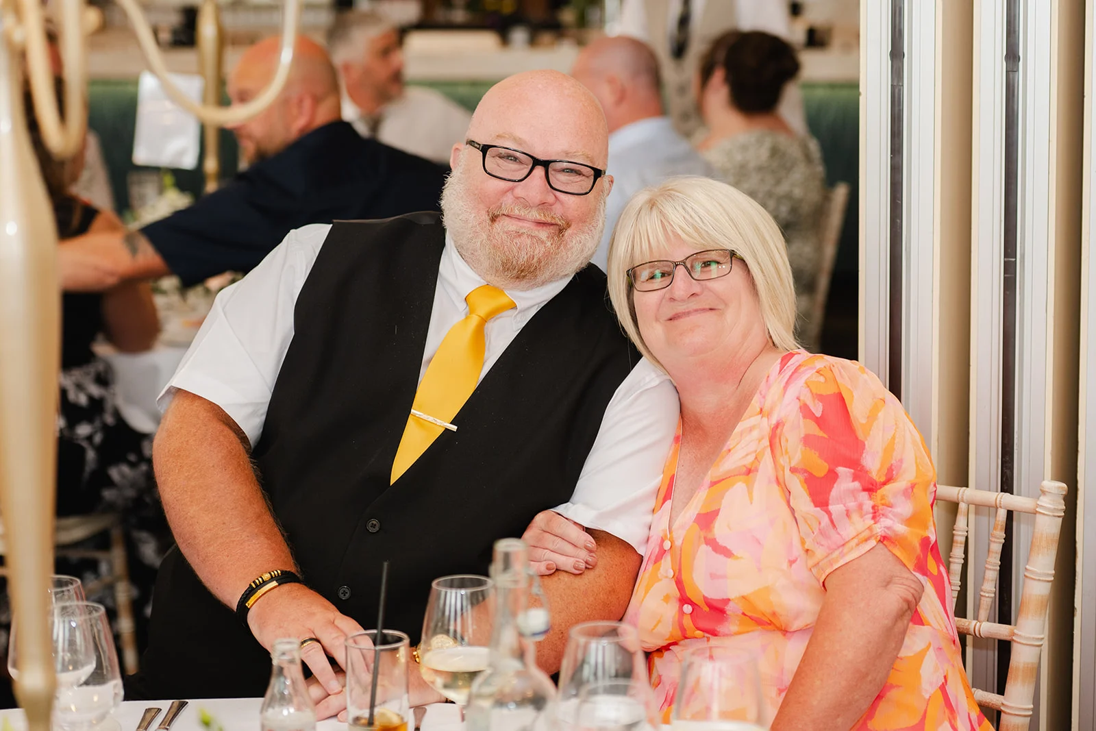 An older couple smiles warmly for the camera seated at a wedding reception table surrounded by guests and wine glasses.