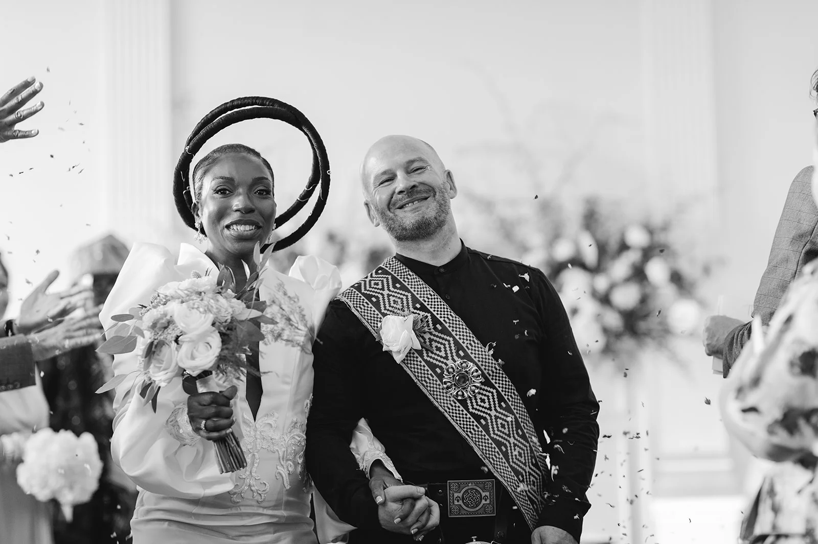 A joyful interracial newlywed couple walk hand in hand through falling confetti, smiling broadly as guests celebrate their marriage.