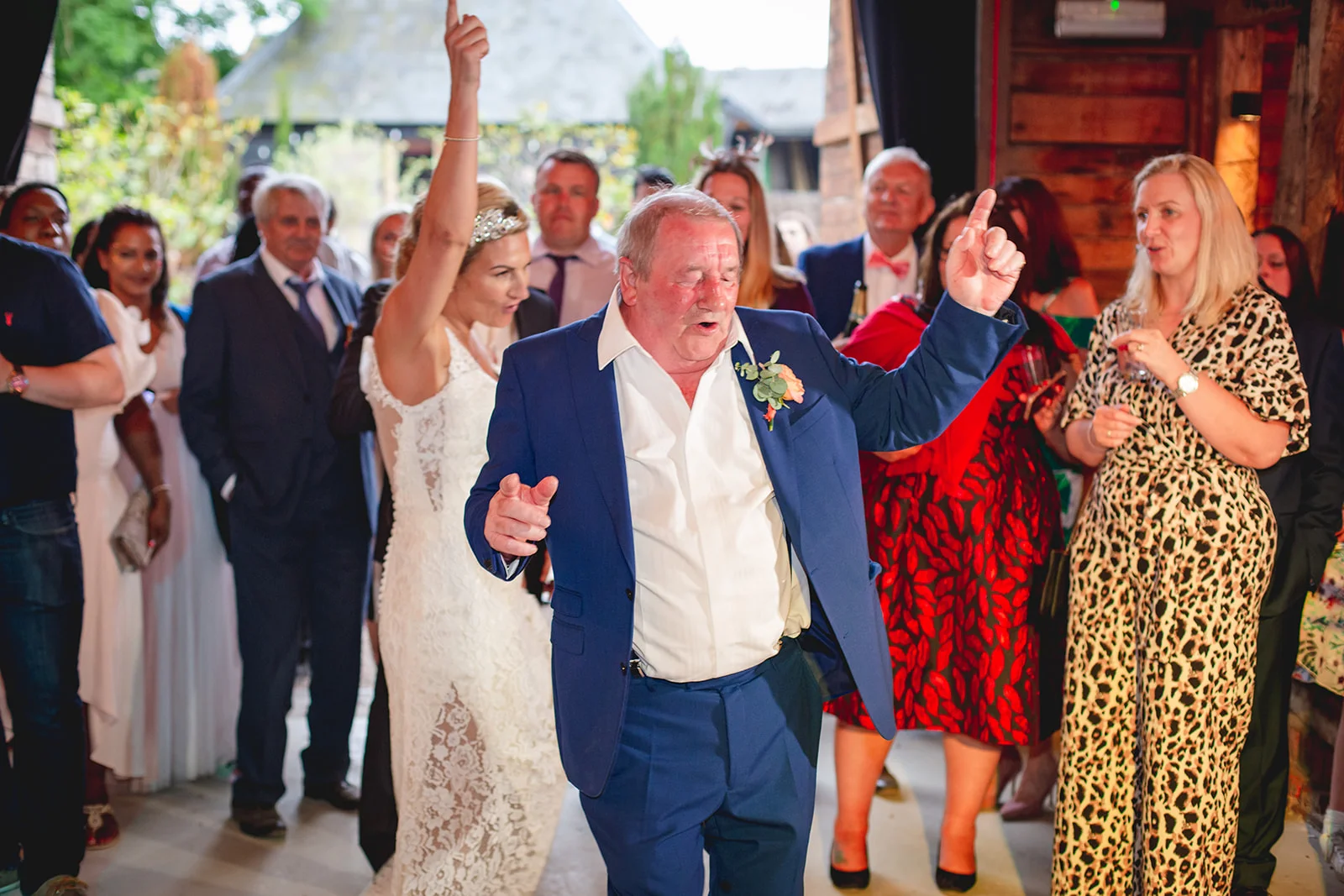 An older man in a blue suit and white shirt dances energetically at a barn wedding reception, surrounded by cheering guests.