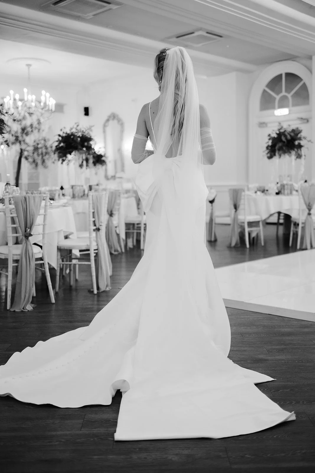A stunning black and white rear portrait of a bride showcasing her elegant wedding gown with a sweeping train, low back, and oversized bow detail inside a grand ballroom reception.