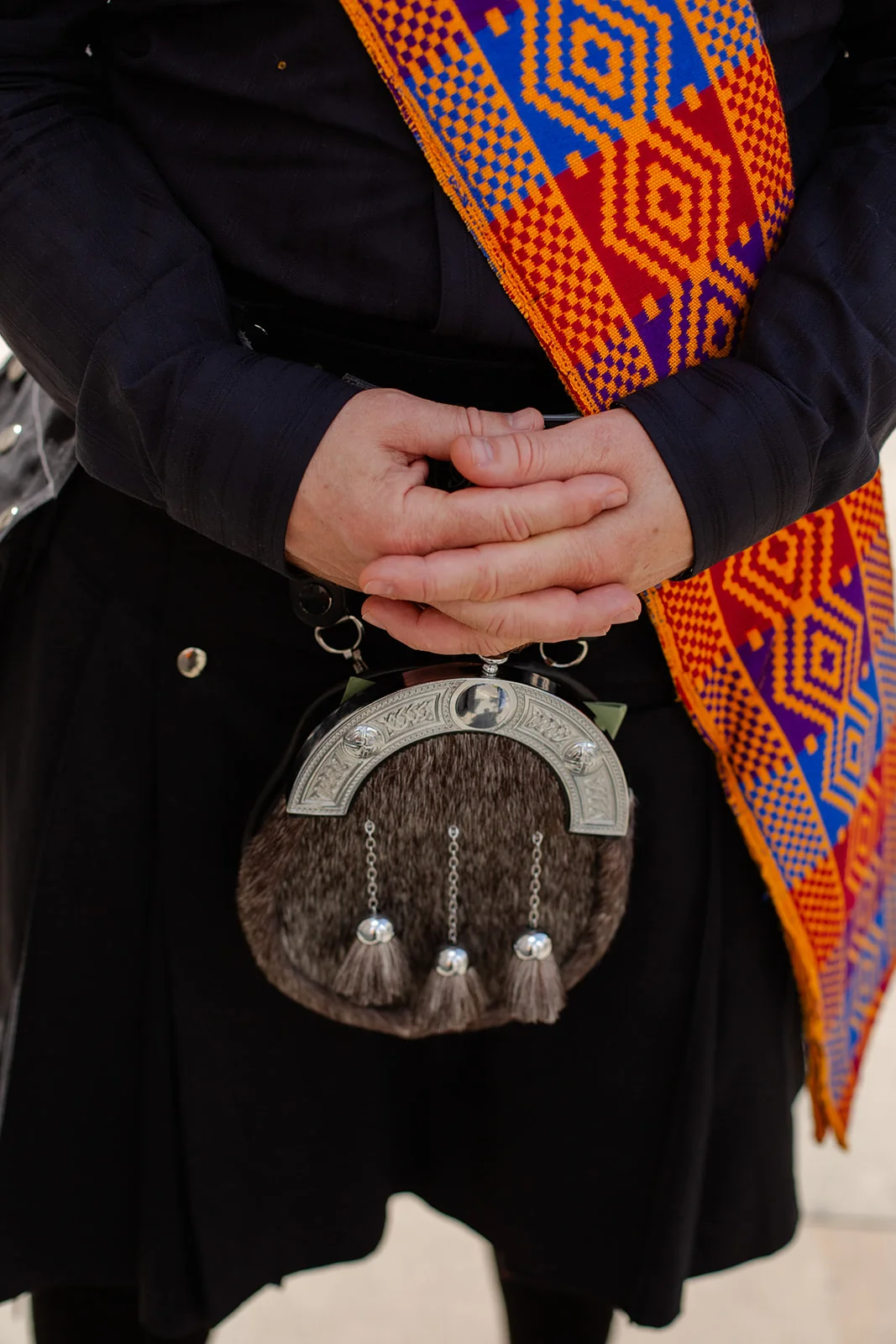 A groom dressed in traditional highland attire, featuring a black kilt, sporran, and a vibrant geometric patterned sash, stands with hands clasped.