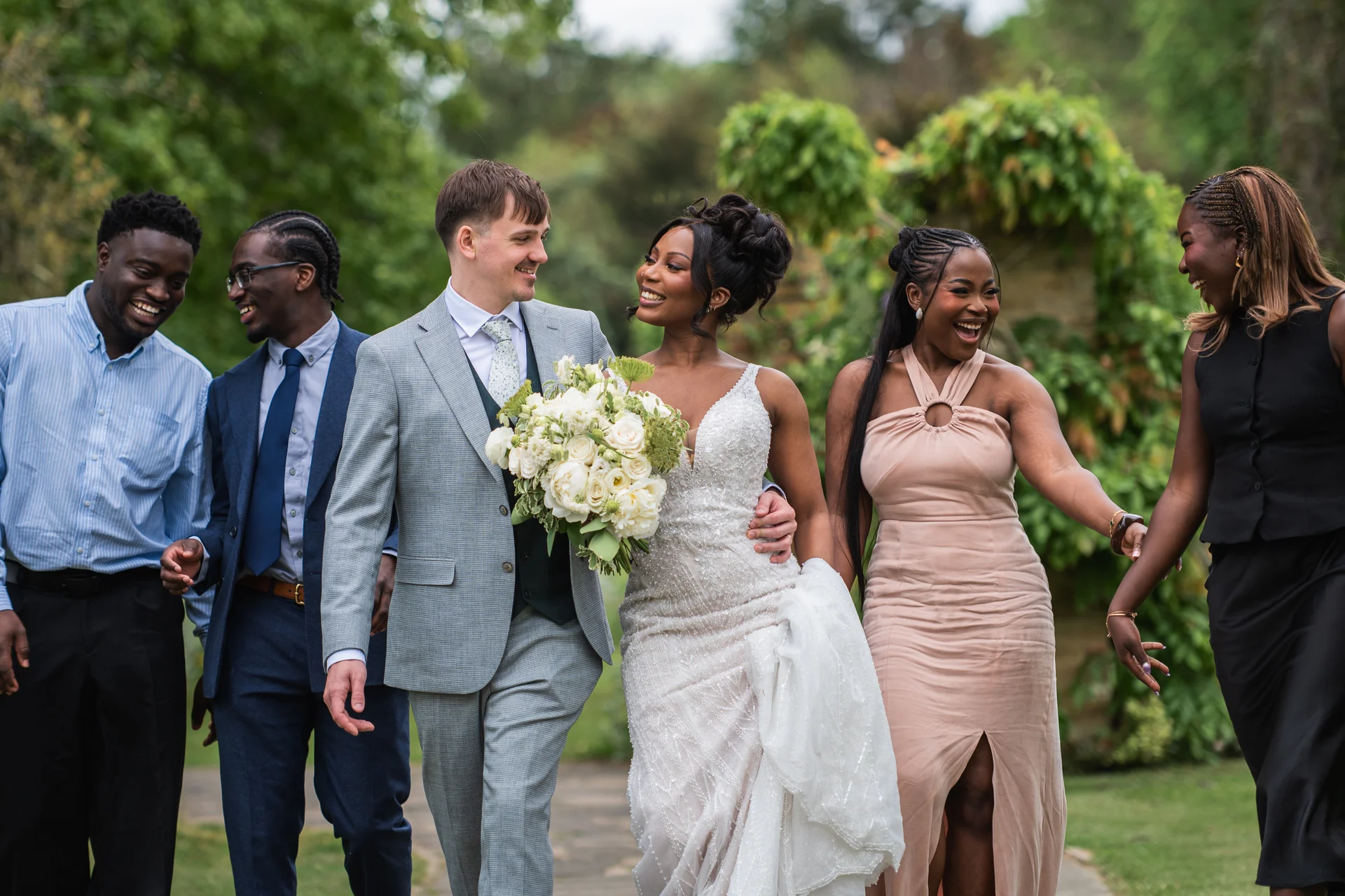 A happy bride and groom walk together outdoors in a lush garden, flanked by laughing groomsmen and bridesmaids in coordinated attire.