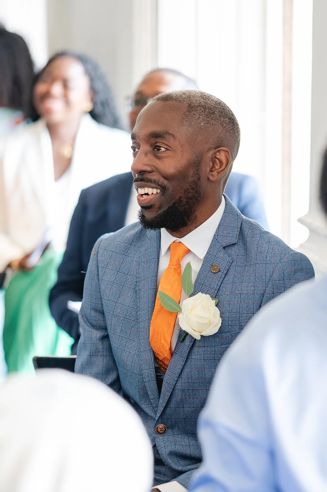 A smiling wedding guest in a checked suit and orange tie with a white rose boutonniere looks off-camera while sitting amongst guests.