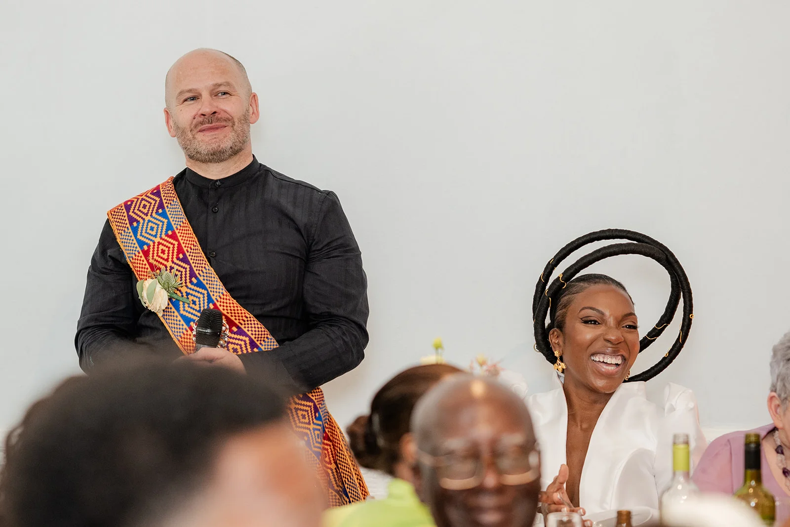 A bald groom in a black shirt with a patterned sash delivers a speech while his Black bride wearing a sculptural halo headpiece laughs joyfully at the wedding reception.