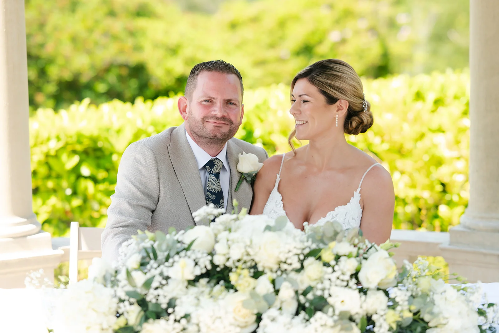 A smiling bride and groom sitting together outdoors under a stone archway behind a lush white floral arrangement during their wedding reception.