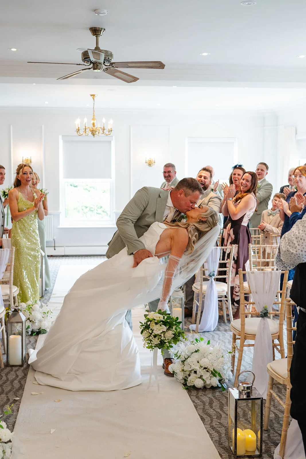 A newlywed couple shares a dramatic dip kiss at the end of the aisle as guests applaud and cheer during their indoor civil ceremony.