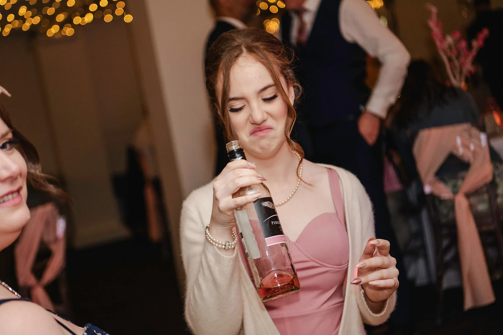 A wedding guest wearing a pink dress and cardigan makes a funny face while inspecting a bottle of wine during the evening reception.