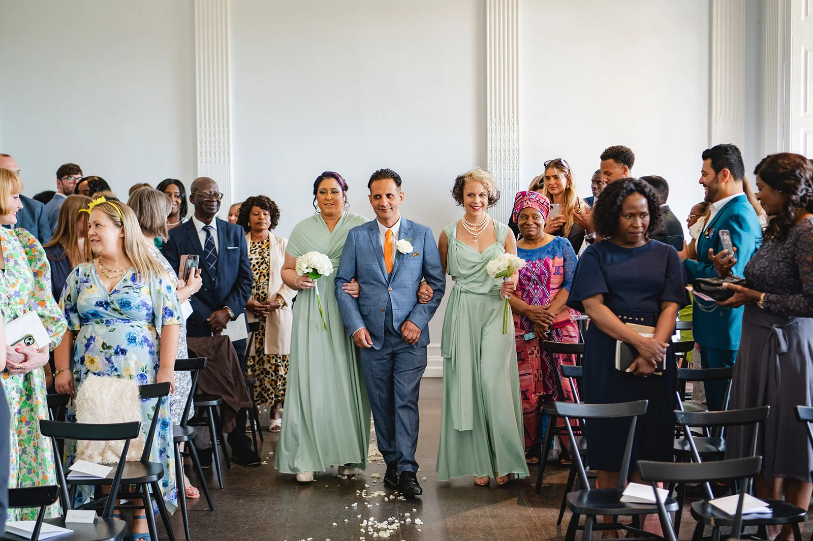 A groom in a blue suit walks down the aisle flanked by two bridesmaids in sage green dresses, holding bouquets while guests watch from seating on either side.