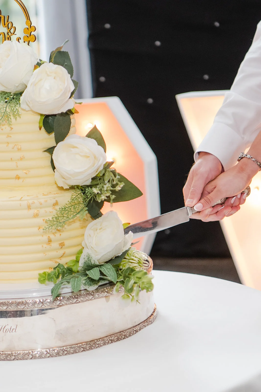 A newlywed couple hold a cake knife together, cutting into a tiered buttercream wedding cake adorned with white roses and gold leaf.