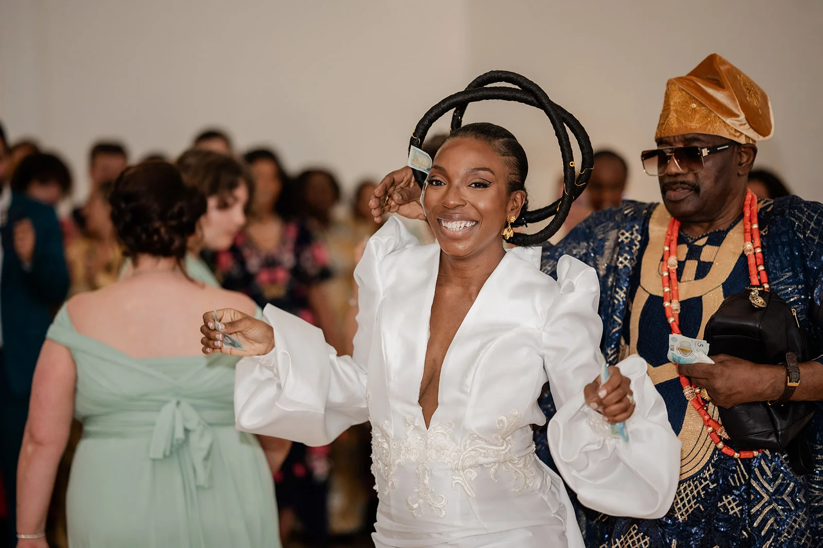 A smiling bride wearing a white dress with balloon sleeves and a sculptural headpiece participates in the money spray tradition during a Nigerian wedding reception.