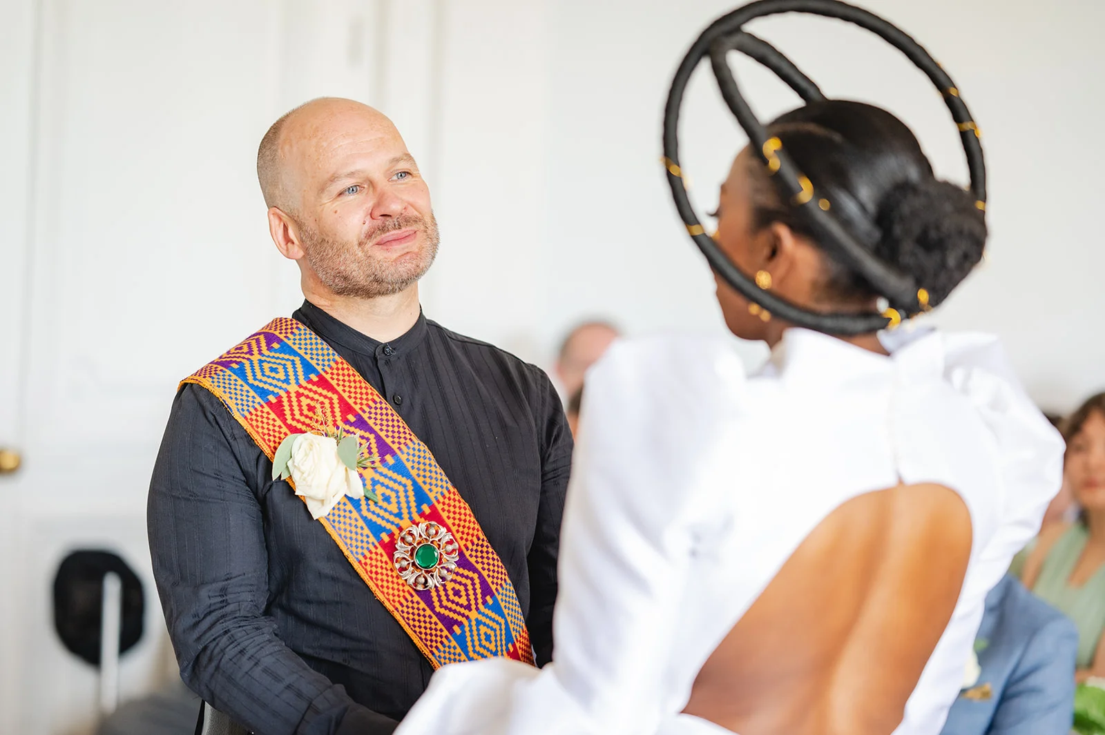 A groom in a black shirt and patterned sash looks adoringly at his bride who wears a striking white open-backed gown and a sculptural circular headpiece during their wedding ceremony.