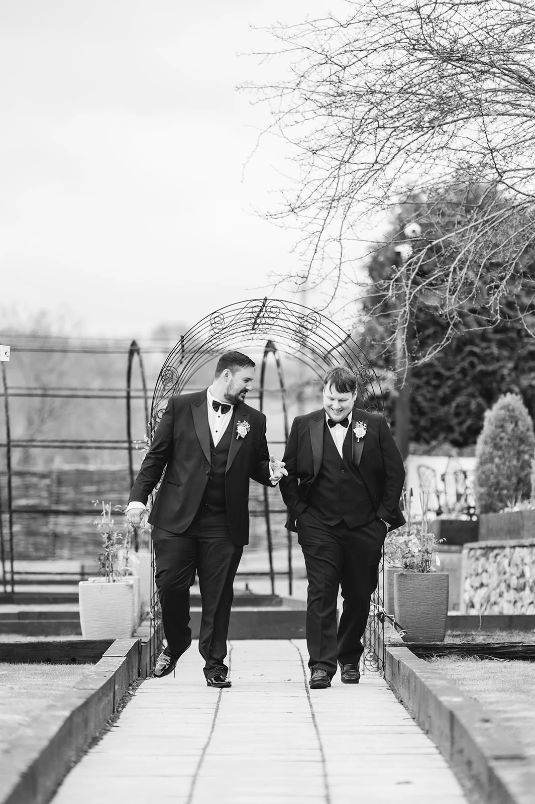 Two grooms in matching black tuxedos smile and walk together along a paved garden path beneath a metal archway.