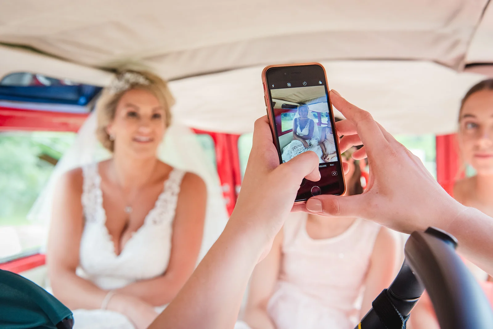 A guest captures a candid photograph of a smiling bride sitting inside a vintage wedding campervan using a smartphone.