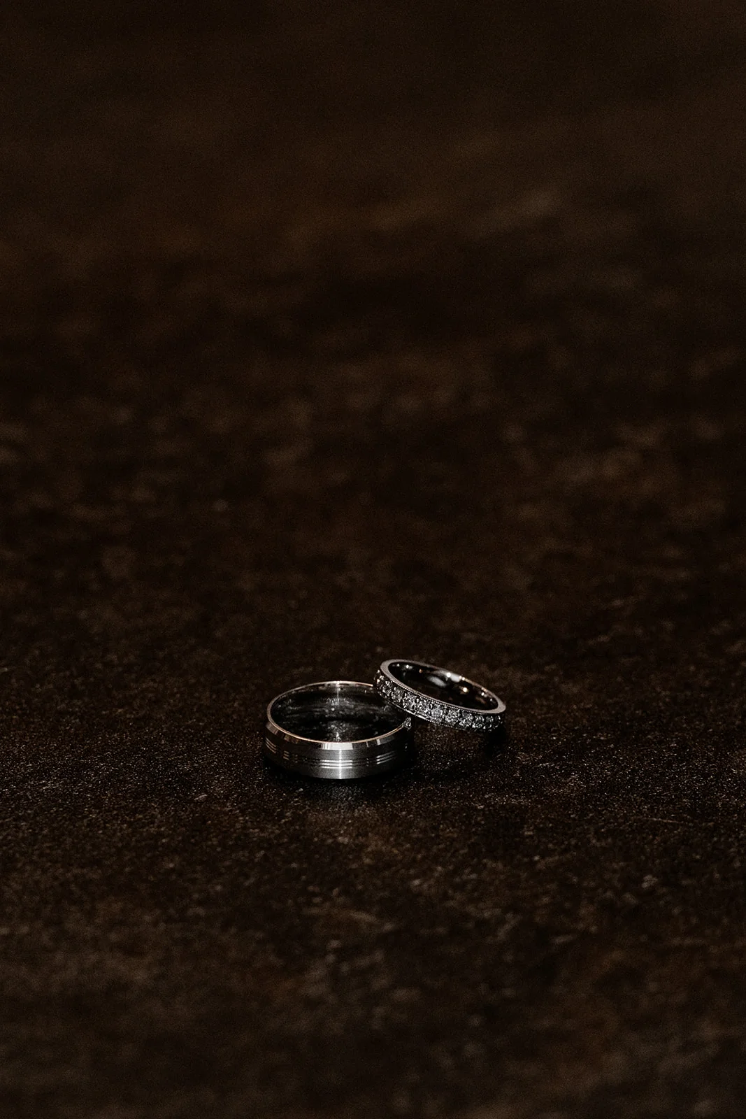 A macro detail shot showcases a pair of elegant wedding rings resting on a dark, textured surface under moody lighting.