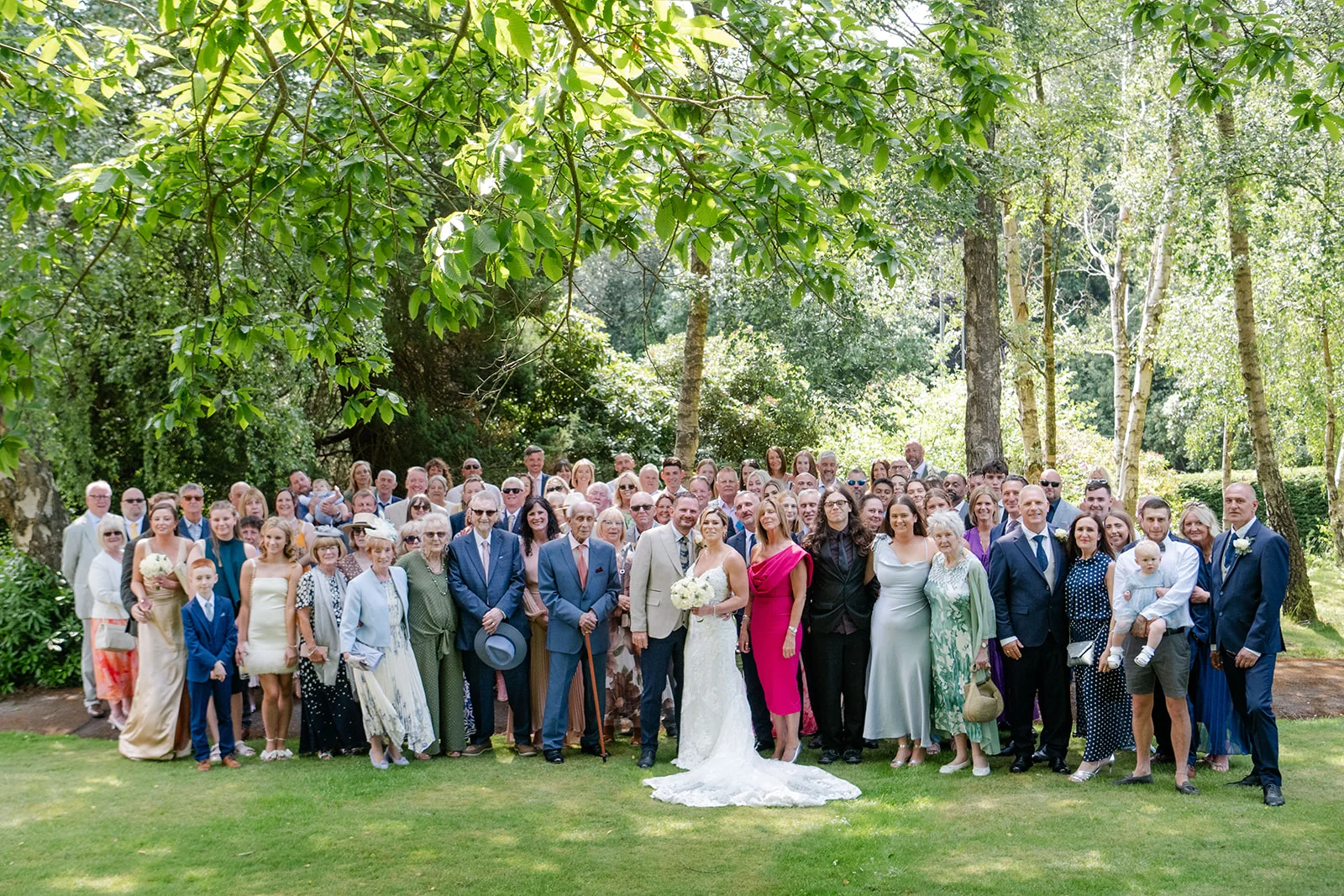 A large wedding party and guests gather for a formal group portrait on a lush lawn under the shade of mature trees during a summer garden wedding.
