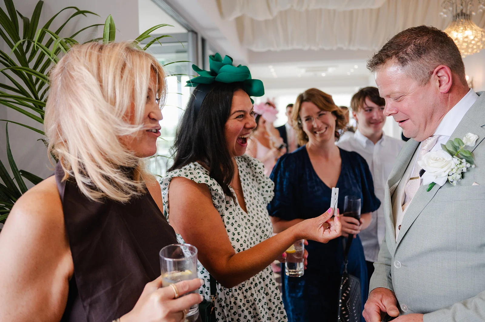 A group of wedding guests laughing and chatting during the drinks reception while a groom in a sage suit looks on.