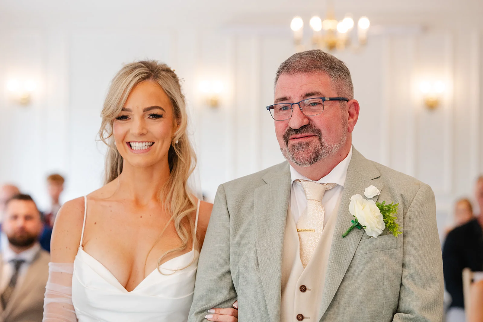 A joyful bride with blonde hair in a satin dress walks down the aisle arm in arm with her smiling father wearing a grey suit and buttonhole.