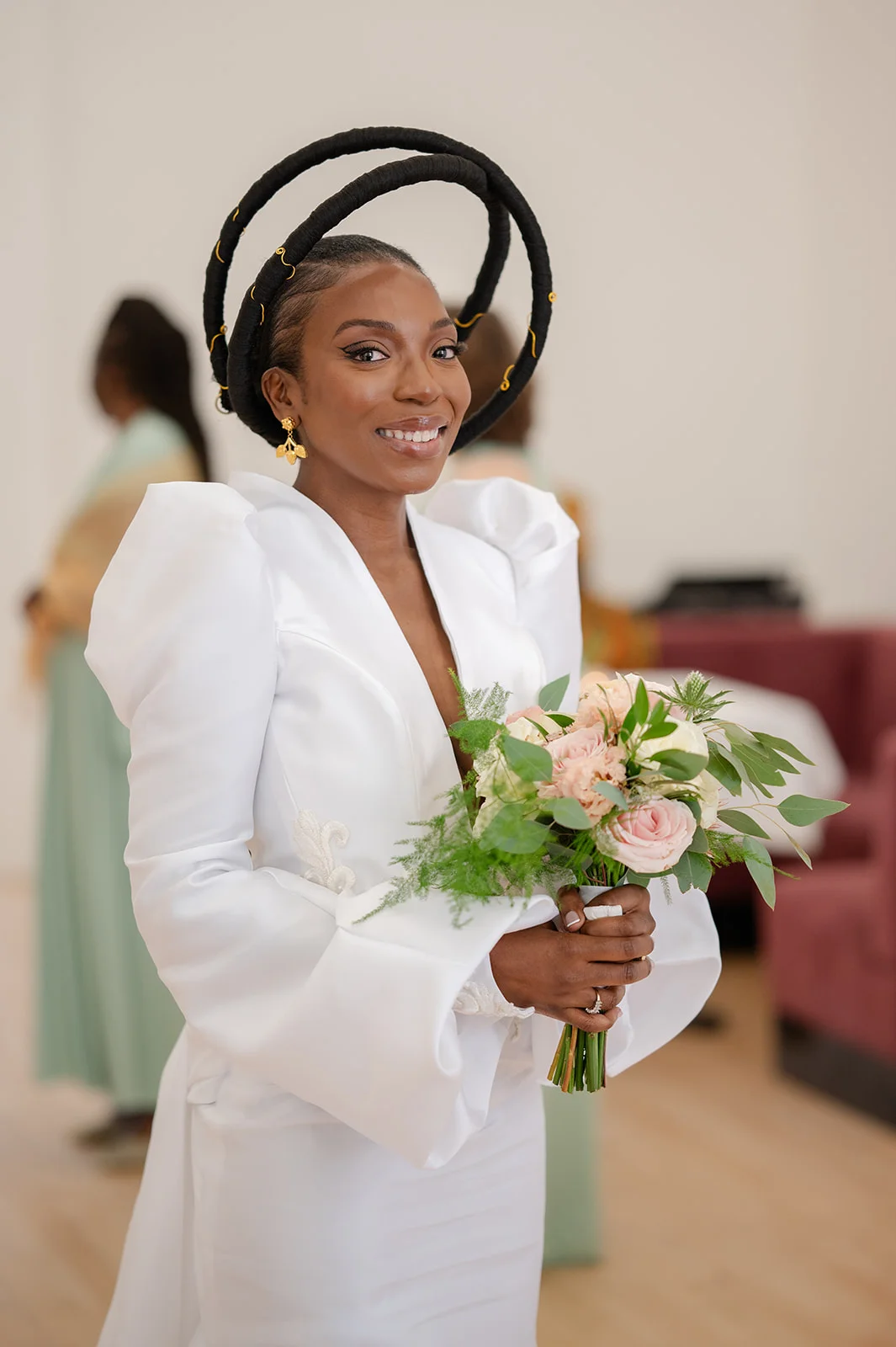 A smiling bride wearing a structured white dress with statement puff sleeves and a unique halo headpiece holds a bouquet of pink roses and greenery.