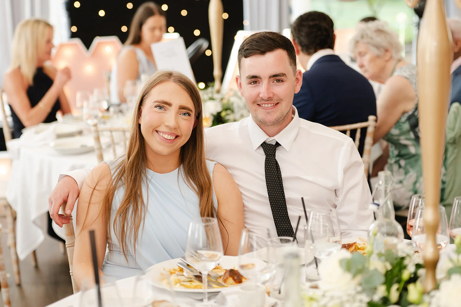 A smiling young couple poses together at a wedding reception table set with white linens, glassware and floral centrepieces.