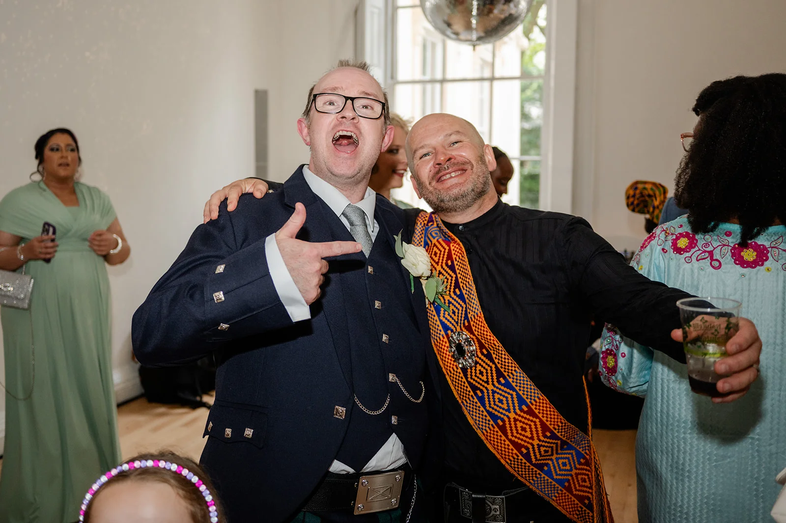 Two smiling grooms in formal wear embrace joyfully during a lively wedding reception celebration.