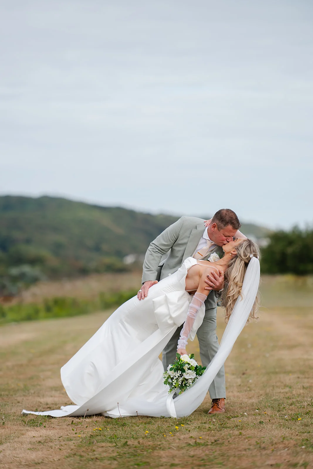 The groom dips the bride in a romantic kiss outdoors against a scenic rolling hills backdrop, with the bride wearing a satin gown and sheer gloves while holding a bouquet.