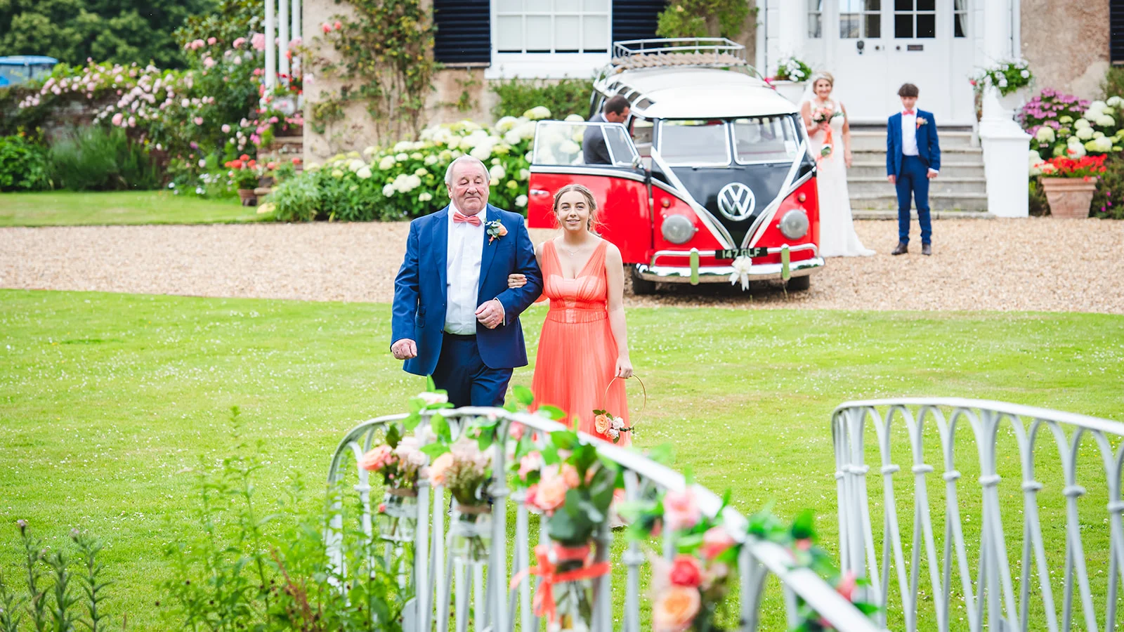 A father of the bride and a bridesmaid walk across a vibrant green lawn towards the camera, with a vintage red and white Volkswagen campervan parked in the background outside a country house.