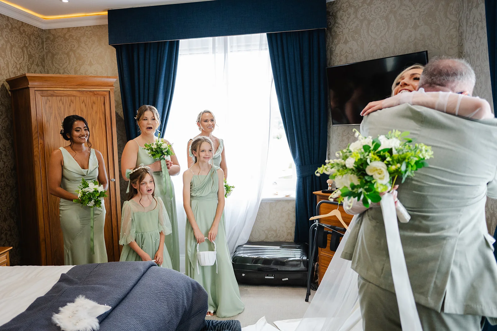 A joyful bride shares an emotional hug with her father in a hotel room while her bridesmaids and flower girls look on with happy expressions.