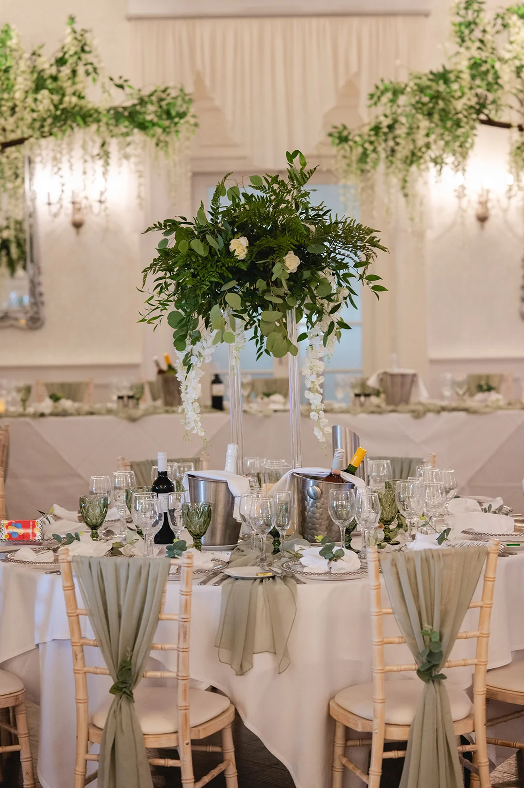 An elegantly styled round wedding table features a tall floral centrepiece with white blooms and greenery, surrounded by sage green glassware and draped chairs.