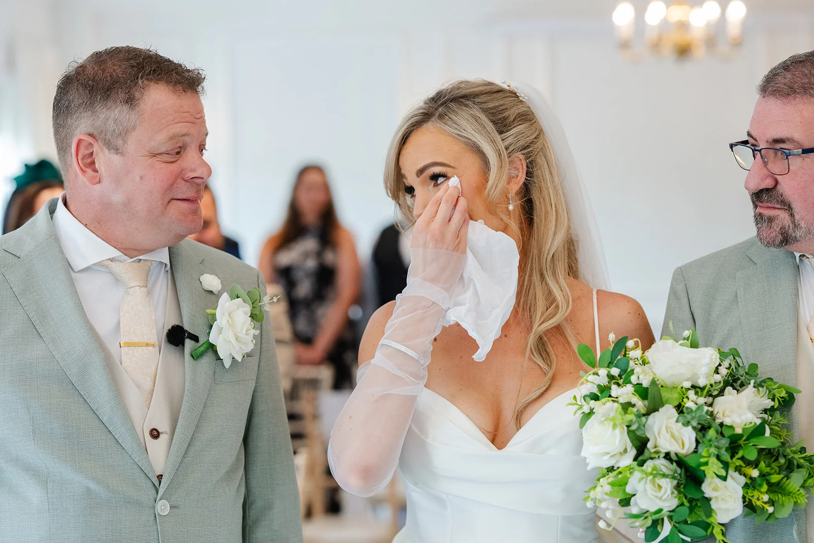 A tearful bride wiping away a happy tear with a tissue while holding a white rose bouquet, flanked by the groom and father of the bride during the wedding ceremony.
