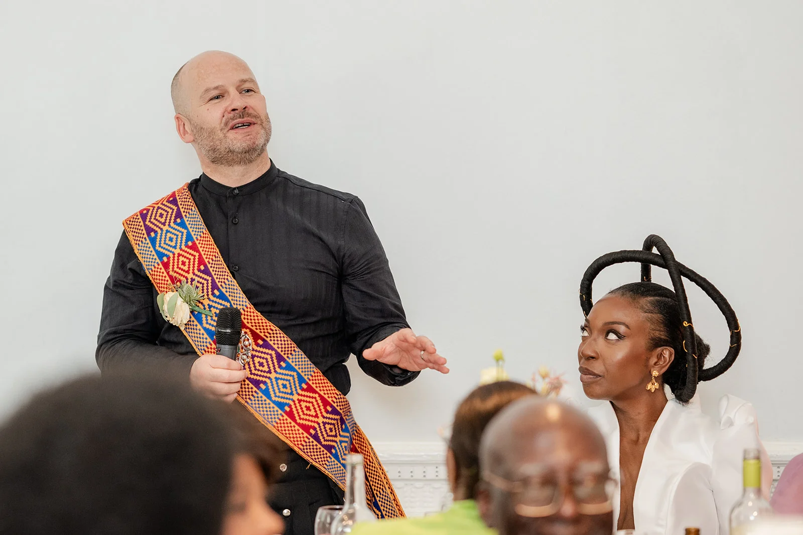 A guest delivers a speech while holding a microphone and wearing a colourful sash, while another guest looks on admiringly with an elaborate headpiece.