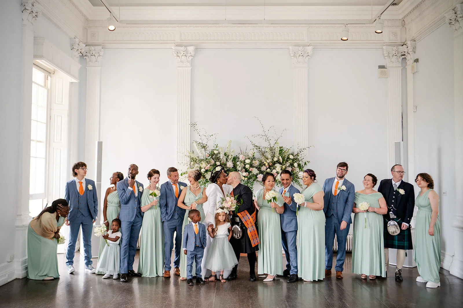 A wide shot of a wedding party standing in a grand hall with sage green and blue attire, as the newlywed couple shares a kiss in front of a floral backdrop.