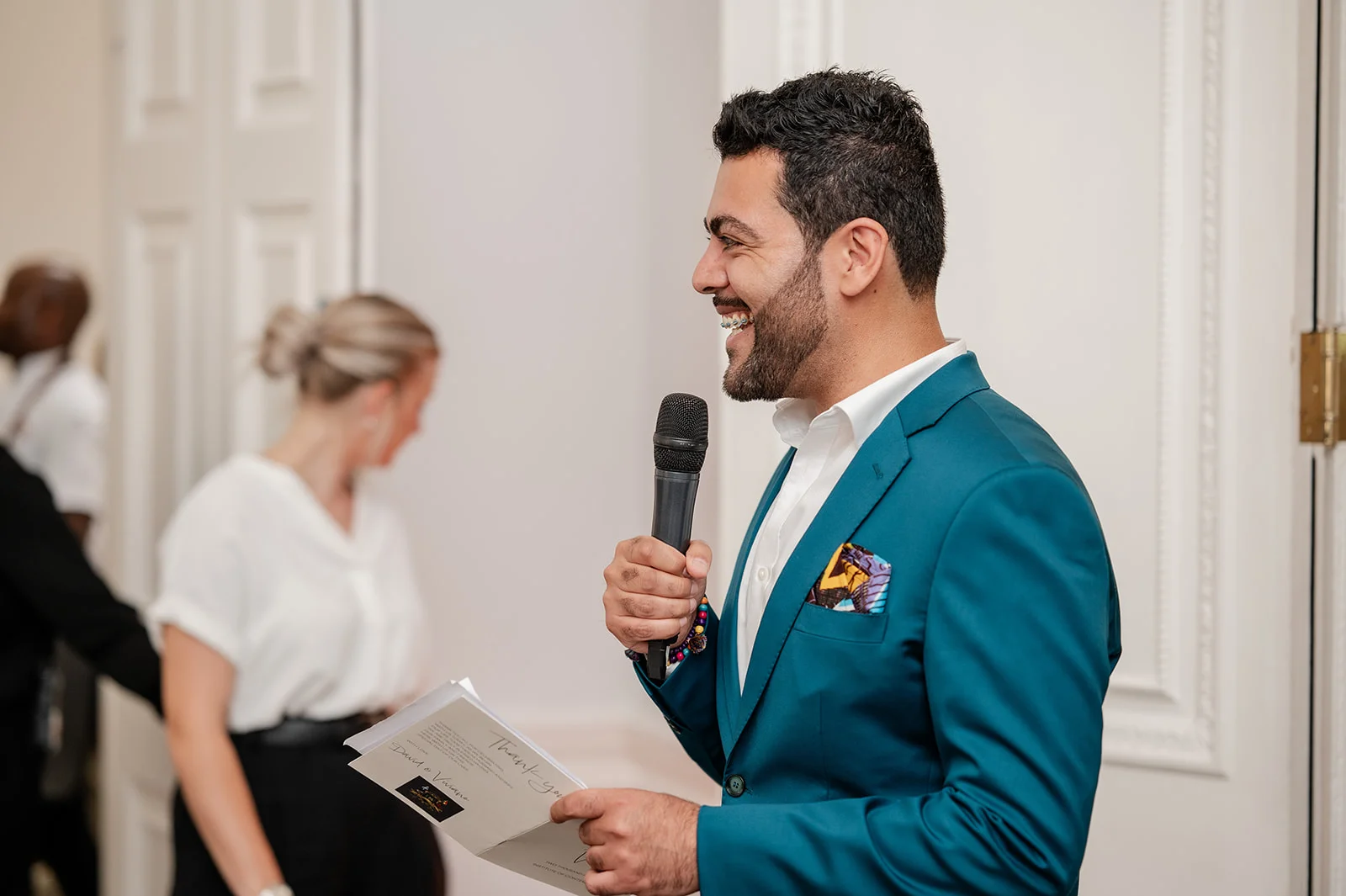 A smiling groom delivers his wedding speech, holding a microphone and a booklet while wearing a vibrant teal suit.