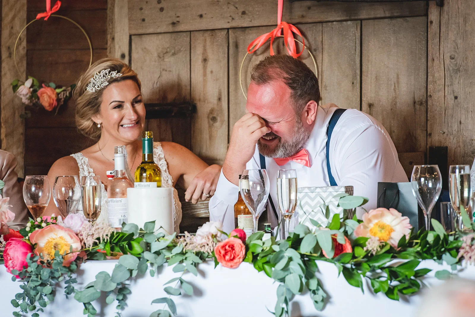 A bride and groom laughing together at the top table during wedding speeches inside a rustic barn venue with a floral garland.