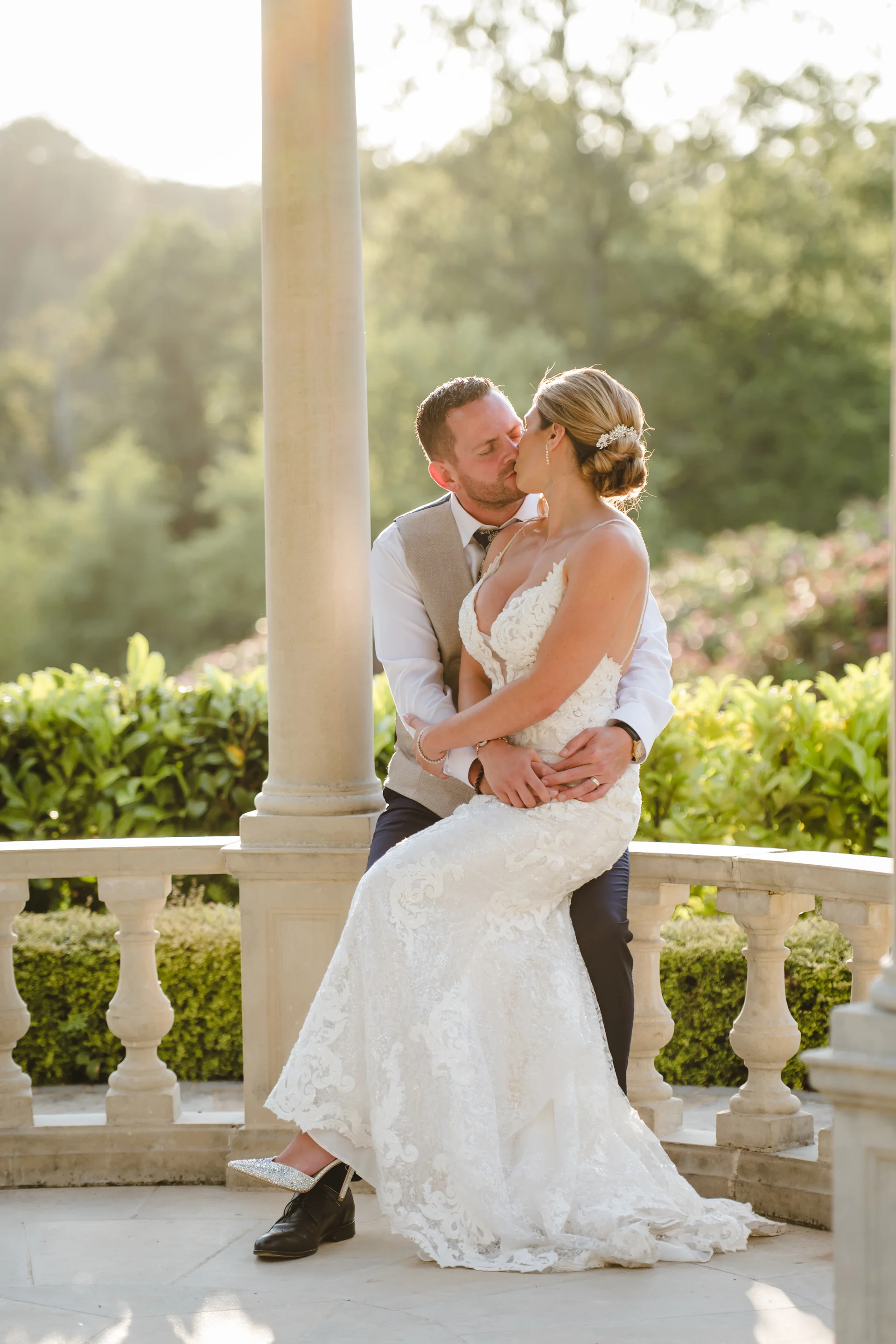 A newlywed couple shares a tender kiss under a stone gazebo during a sunlit golden hour portrait session in the gardens.