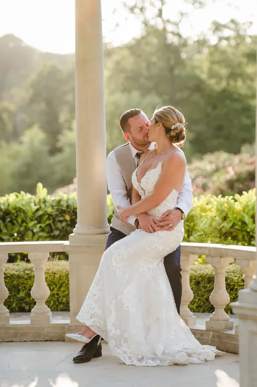 A newlywed couple shares a tender kiss under a stone gazebo during a sunlit golden hour portrait session in the gardens.