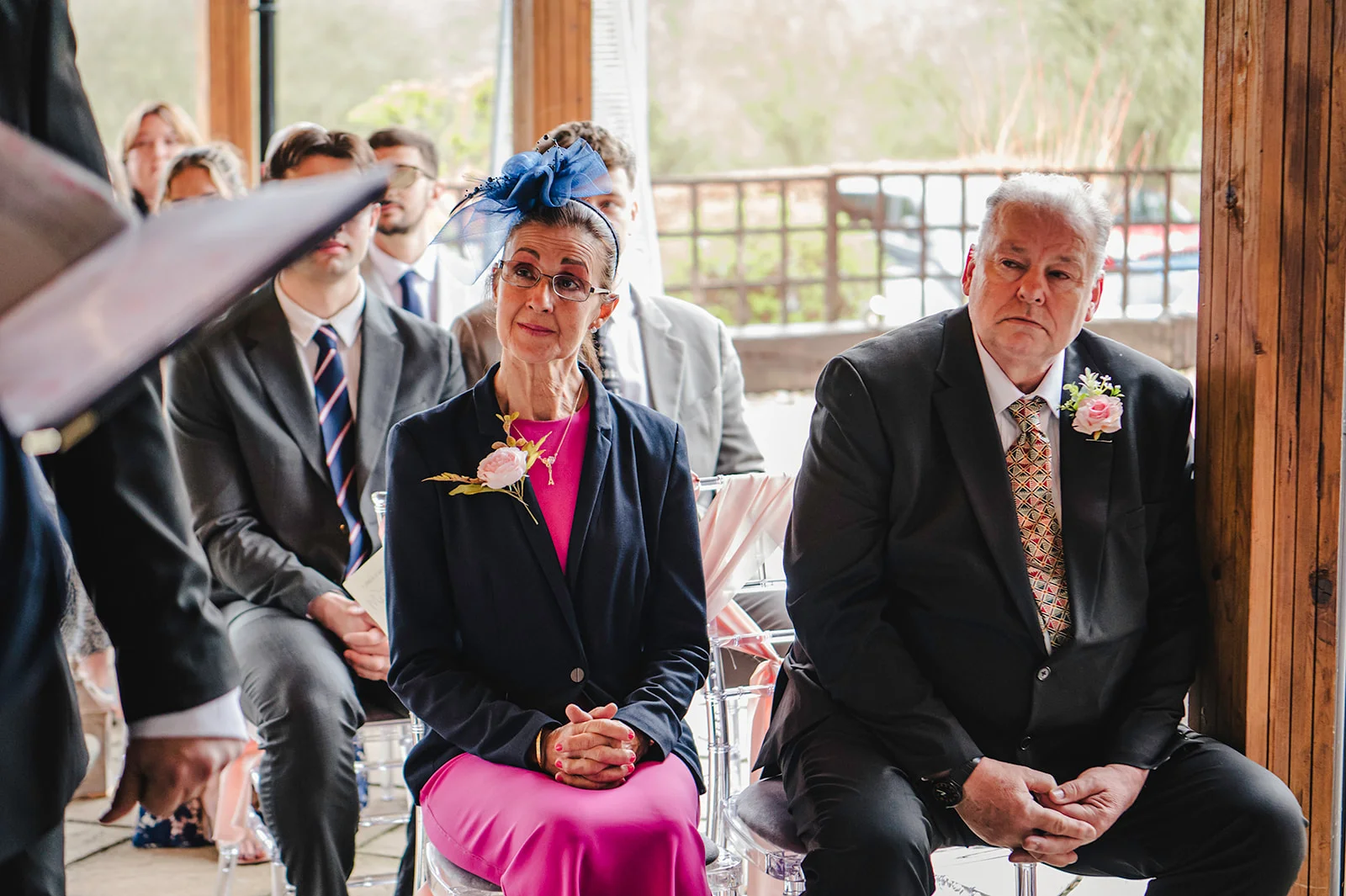 The mother and father of the bride sit attentively amongst seated guests during a wedding ceremony.