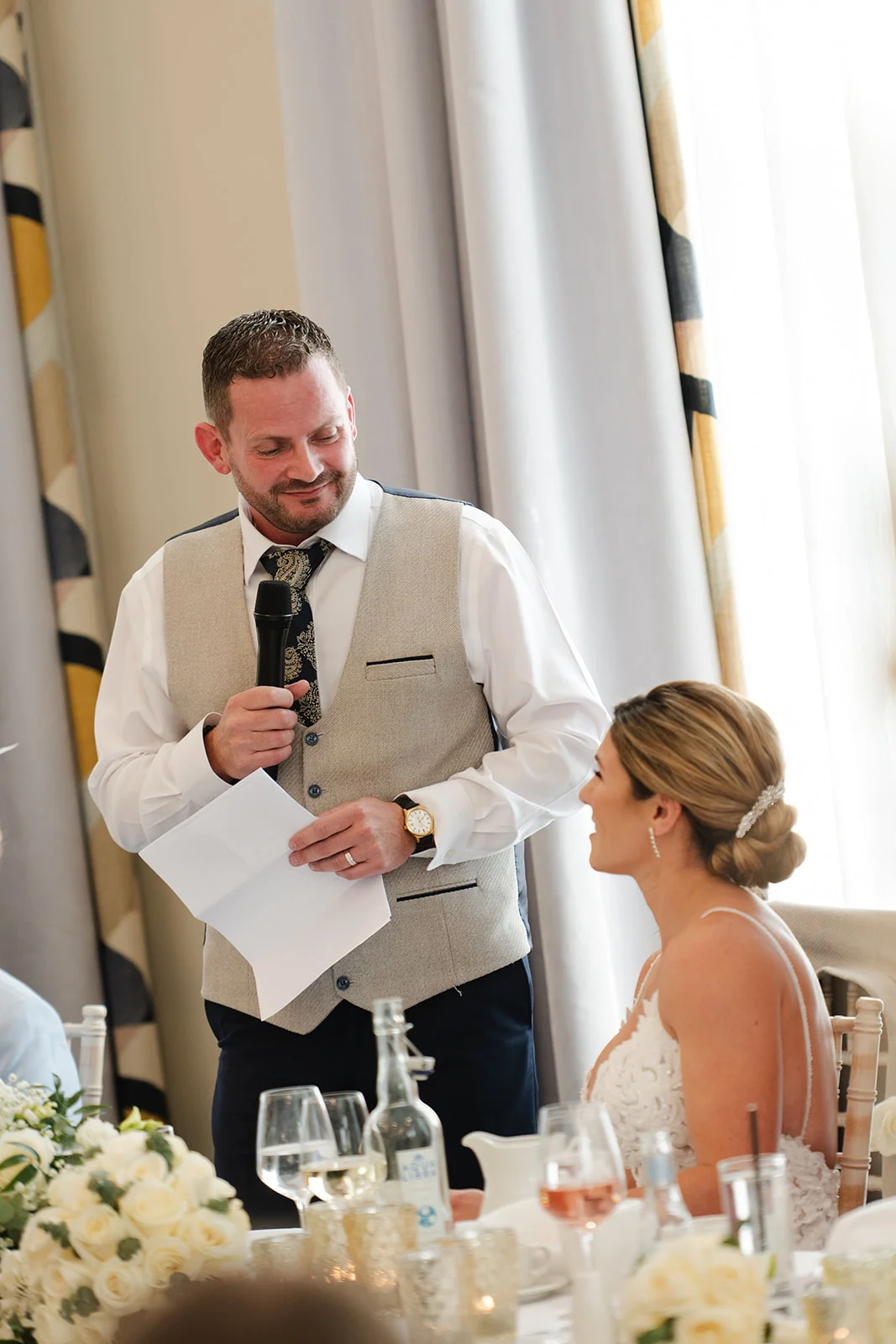 A groom gives a heartfelt speech holding notes and a microphone, while the bride looks on with a smile at the wedding reception table.