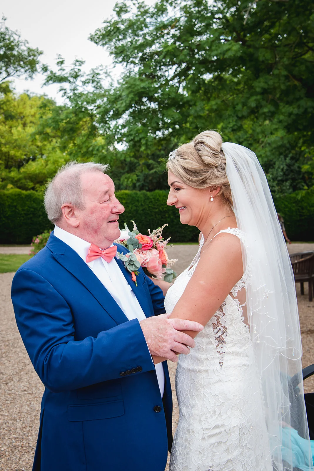 A proud father shares a joyful, emotional moment with his smiling daughter in her lace wedding dress and veil outdoors in a garden.