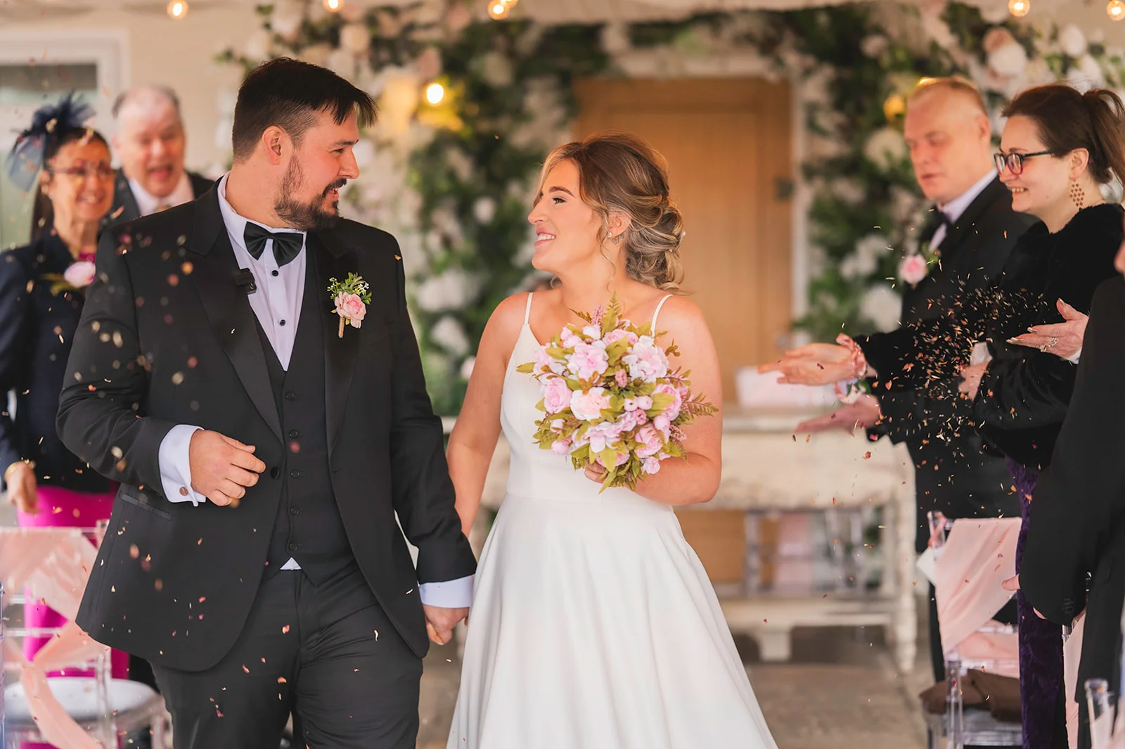 Newlyweds walk hand in hand down the aisle smiling at each other as family and guests shower them with confetti under a floral arch.