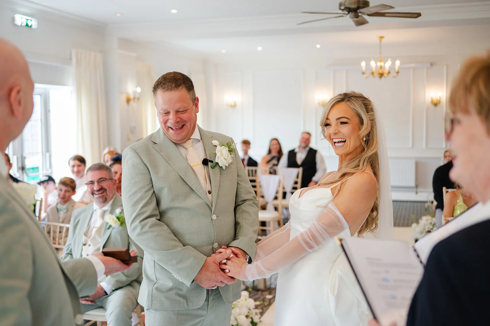 A smiling bride and groom hold hands and laugh during their bright indoor civil ceremony, surrounded by seated guests.