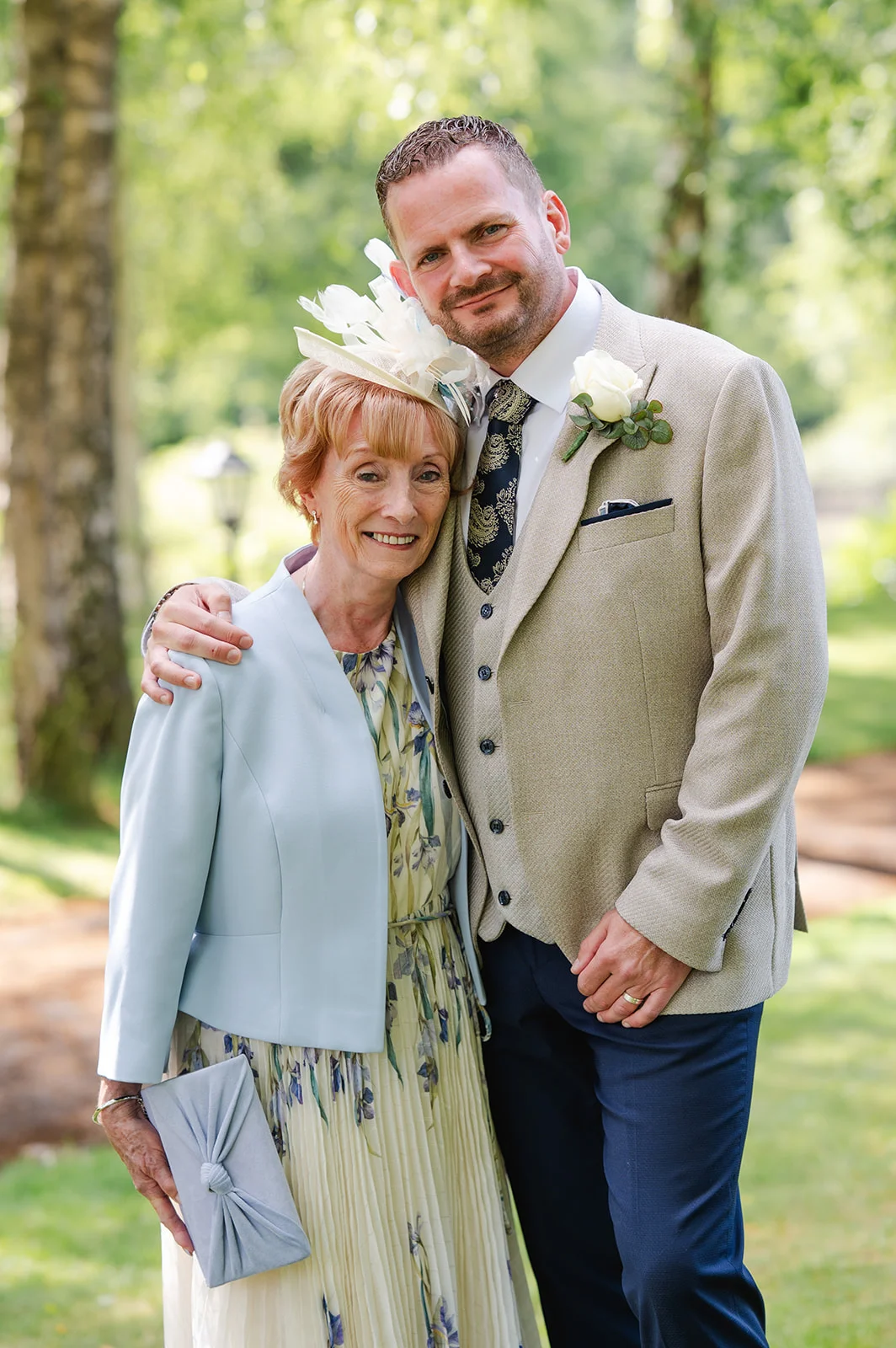 A groom poses outdoors with his mother, who wears a pale blue jacket and matching fascinator, while standing in a sunlit garden setting.