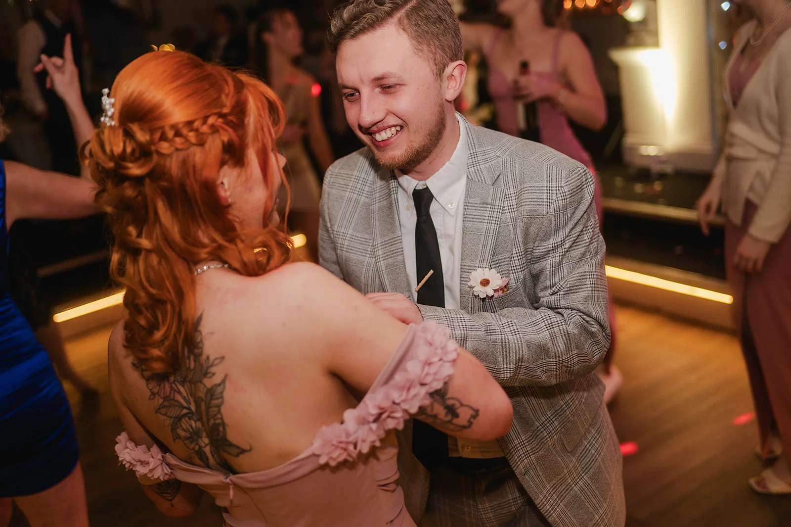A smiling groom in a grey checked suit and black tie dances joyfully with his red-haired bride on the evening reception dance floor.