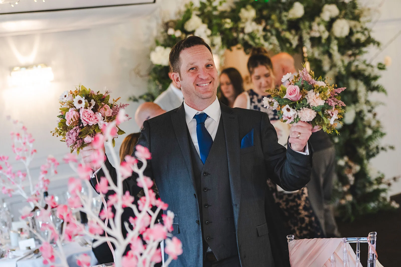 A smiling groom wearing a charcoal three-piece suit and blue tie holds up two floral bouquets during a wedding reception.